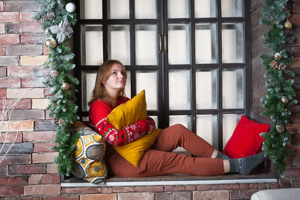 Una chica rubia en un suéter rojo se sienta en el alféizar de la ventana. — Foto de Stock