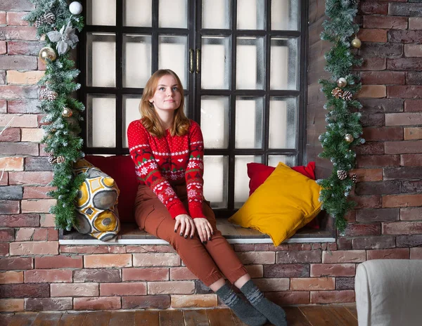 Een blond meisje in een rode trui zit op de vensterbank. — Stockfoto