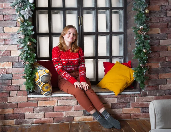 Een blond meisje in een rode trui zit op de vensterbank. — Stockfoto