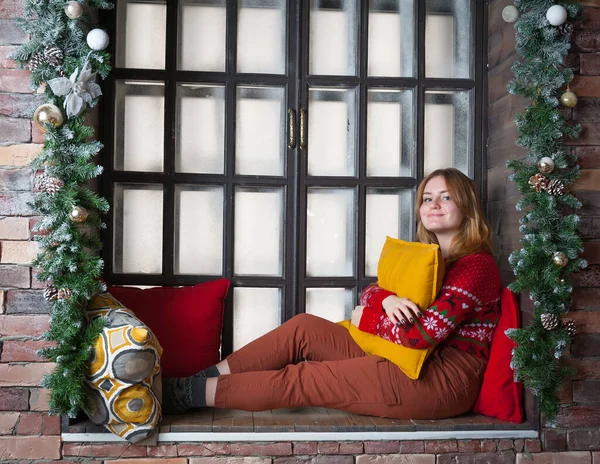 Een blond meisje in een rode trui zit op de vensterbank. — Stockfoto