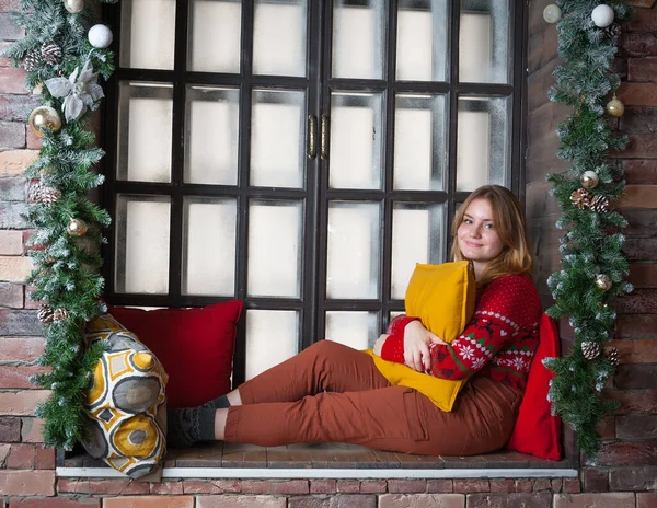 Een blond meisje in een rode trui zit op de vensterbank. — Stockfoto