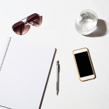 Sunglasses, water, phone and notepad flat lay on a white table