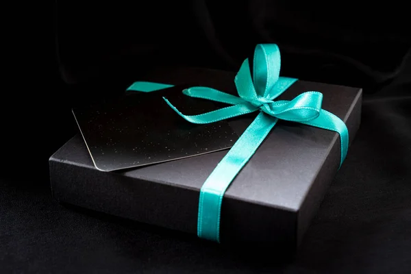 Siyah arkaplanda yazılı bir kartın olduğu turkuaz bir kurdeleyle bağlanmış kara kutu.