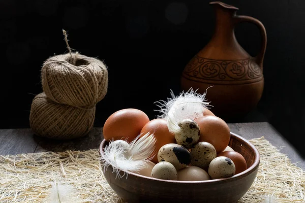 Kil bir kasede tavuk ve bıldırcın yumurtaları. Koyu ahşap bir masada, arka planda vrevka skeins ve kilden bir sürahi var.