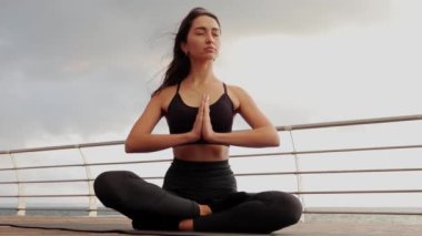 Gün batımında kumsalda bir kayanın üzerinde oturmuş yoga yapan genç bir kadın silueti..