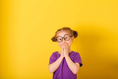 Little cute caucasian funny surprised happy kid child baby girl with glasses in purple T-shirt on yellow wall background. children studio portrait. girl presses her hands to her mouth