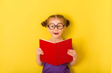 Küçük kız kitap kitabı tutuyor ve şaşırdı. Sarı arka planda sevimli, komik, neşeli, neşeli bir çocuk. çocuk stüdyo resmi.