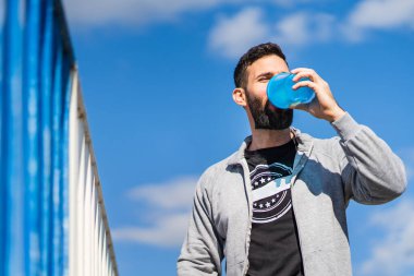 Yetişkin erkek, beyaz atlet elinde bir şişe takviye içkisiyle dışarıda duruyor. Güneşli bir günde içiyor. Ön manzara, belden yukarı fotokopi alanı.
