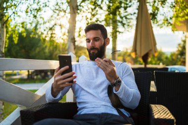 Güneşli bir sonbahar gününde kafede cep telefonu kullanarak sakallı, yetişkin ve beyaz bir adamın portresi. Boş zamanların konsepti uzay kopyası.