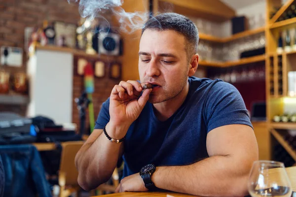 一人でテーブルに座っている若い大人の白人男性家やレストランで葉巻を持っている喫煙彼の頭の周りに煙を見下ろす日にTシャツを着て — ストック写真