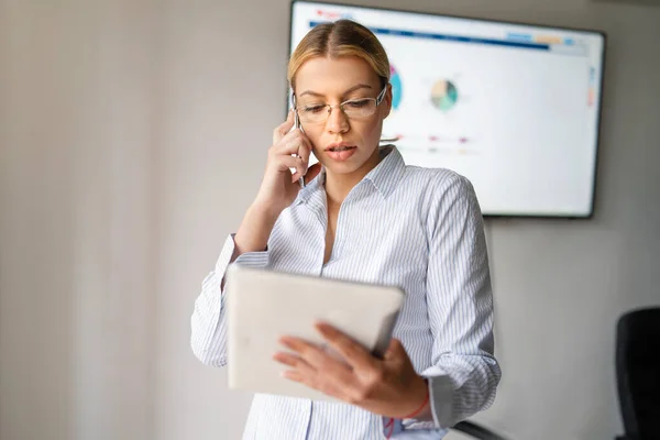Portrait of young beautiful caucasian woman working at office. Blonde girl standing making a call. Front view of woman using phone at the office reading report. business and female empowerment concept