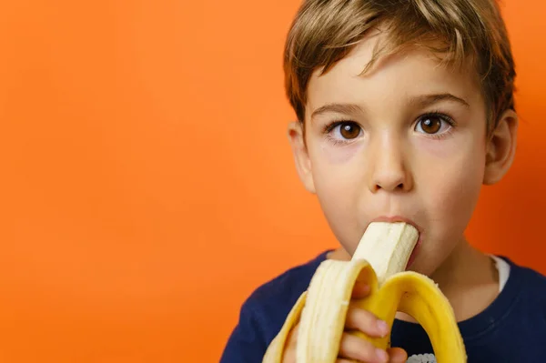 Small caucasian boy eating banana in front of orange background wall