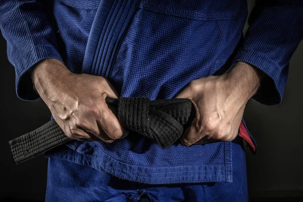 Kimliği belirsiz beyaz adamın elinde Brezilya jiu jitsu bjj siyah kuşağı tutarken karanlıkta kimono gi giydiği dövüş sanatları ustalığı ve güven kazanma konsepti.