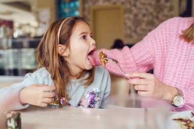 Annesi elinde çatal ve yiyecekle esmer kızını beslerken lokantada masada oturan ağzı açık küçük beyaz bir kızın portresi.