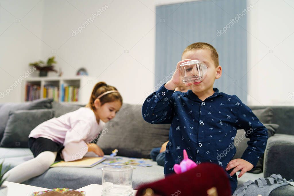 Small four years old caucasian boy holding glass drinking water at home in front of the sofa bed while his sister girl is playing