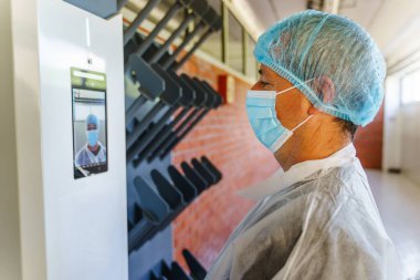 Side view of caucasian man doctor scientist wearing protective gear standing in front of electronic face recognition terminal with digital temperature control camera checking in before going to work clipart