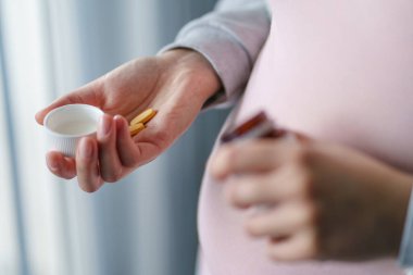 Meçhul hamile beyaz kadının karnını kapatın. Elinde şişeyle evde ilaç veya vitamin takviyesi alıyor. Hamilelik ve takviye konsepti fotokopi alanı.
