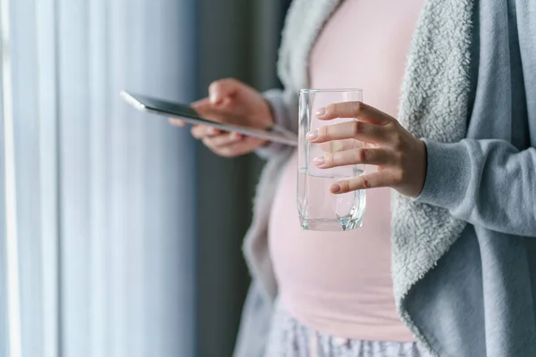 未知の原因不明の女性の手に閉じるU妊娠中の母親が窓のそばに立っている間 水とデジタルタブレットのガラスを保持 妊娠と飲料水の概念 — ストック写真