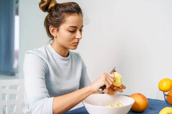 Caucasian young woman peeling fruit while sitting by the table at home - side view on pretty girl holding knife and apple preparing meal - healthy eating concept copy space