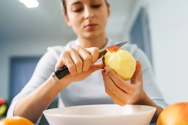 Caucasian young woman peeling fruit while sitting by the table at home - front view on pretty girl holding knife and apple preparing meal - healthy eating concept copy space