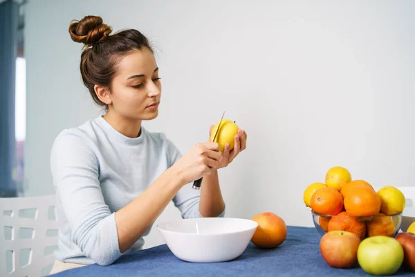 Caucasian young woman peeling fruit while sitting by the table at home - side view on pretty girl holding knife and apple preparing meal - healthy eating concept copy space