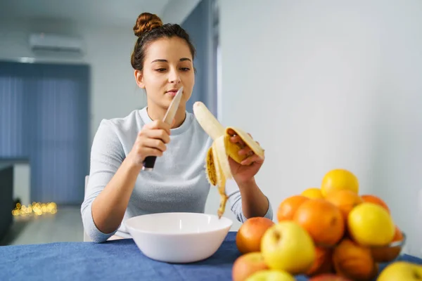 Caucasian young woman peeling fruit while sitting by the table at home - front view on pretty girl holding knife and banana preparing meal - healthy eating concept copy space