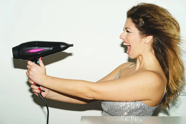 Side view portrait of adult caucasian woman holding electric hair dryer in front of her face - Beautiful woman drying hair and screaming exited in front of white wall - Hair style beauty concept