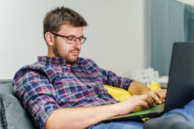 Evinde çalışan genç girişimci bilgisayarla rapor alıyor - beyaz bir adam evinde kanepede otururken dizüstü bilgisayarla yazı yazıyor - evdeki serbest yazar kavramının uzay çalışmalarını kopyalıyor