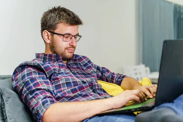 Evinde çalışan genç girişimci bilgisayarla rapor alıyor - beyaz bir adam evinde kanepede otururken dizüstü bilgisayarla yazı yazıyor - evdeki serbest yazar kavramının uzay çalışmalarını kopyalıyor