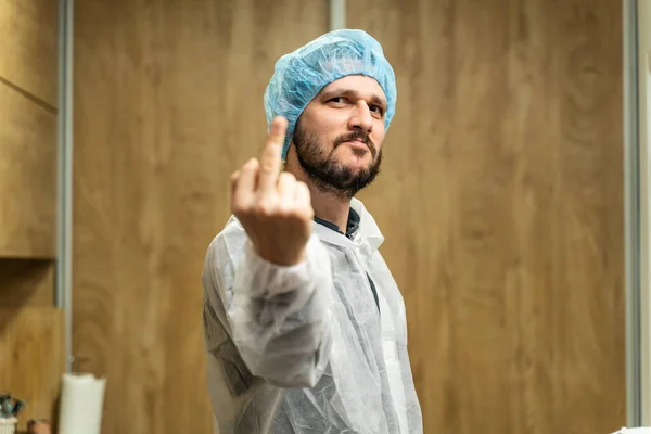 Portrait of man in bouffant cap and protective equipment medic doctor or worker standing in front of wooden wall indoor looking to the camera making gesture showing middle finger bad attitude concept