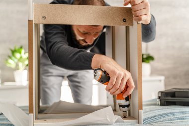 Tek başına sakallı bir adam elinde elektrikli tornavidayla ev eşyaları topluyor ve talimatlara bakıyor.