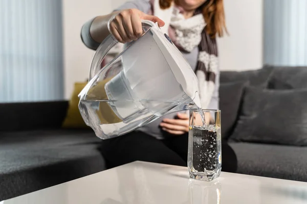 Unknown woman pouring filtered water in glass on the table from the plastic filter jug at home in day - clean water hydration healthy living concept