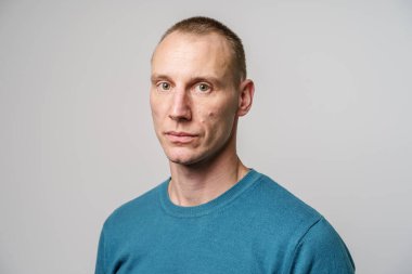 Stüdyo, beyaz duvarın önünde kısa sarı saçlı, mavi kazaklı, yetişkin, ciddi beyaz bir adamın portresi. 35 yaşında.