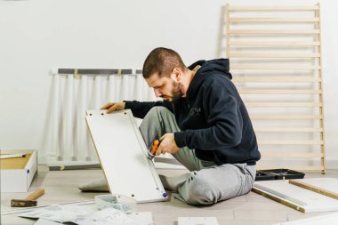 Tek başına sakallı bir adam evde elektrikli tornavida ve kontraplak parçaları taşıyan kişisel montaj mobilyalarını birleştiriyor. Yarı uzunlukta ön manzara DIY konsepti. Gerçek insanlar boşluğu kopyalıyor.