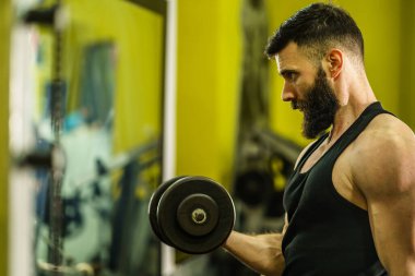 Spor salonunda vücut geliştirme eğitimi alan beyaz erkek sporcunun portresi. Koyu renk gömlekli, siyah saçlı ve sakallı halterli halterler giyiyor. Fotokopi alanı kaldırıyor.