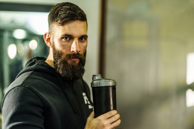 Siyah kapüşonlu genç beyaz erkek sporcunun ön manzara portresi. Elinde protein takviyesi ile spor salonunda duran siyah saç ve sakal fotokopi alanı.