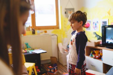 Dört yaşında küçük beyaz bir çocuğun portresi. Evde, dağınık bir odada pijama giymiş oyuncaklarla çevrili bir şekilde duruyor. Çocukluk aile kavramı.