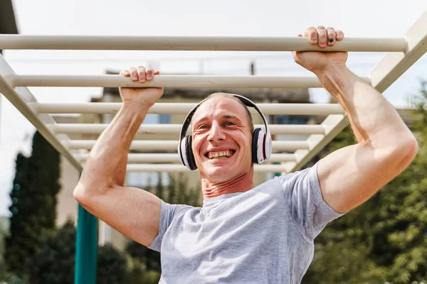 Fitness male athlete smiling while exercising on monkey bars. Cross-fit man working out arms swinging on brachiation ladder as strength training routine in sunny day outdoor strength fitness concept