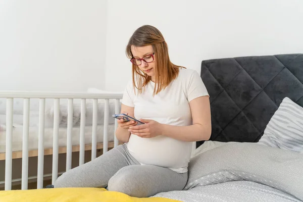 ベッドルームでベッドに座っている間に携帯電話を使用して妊娠中の白人女性 明るい部屋で妊娠中にテキストメッセージやインターネットの閲覧を送信する女性 妊産婦の概念 — ストック写真