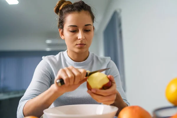 Caucasian young woman peeling fruit while sitting by the table at home - side view on pretty girl holding knife and apple preparing meal - healthy eating concept copy space