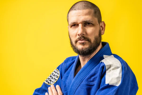 Portrait of adult man in blue gi kimono standing in front of yellow background with copy space looking to the camera