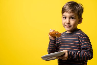 Kafkasyalı küçük bir çocuğun ön görüntüsü sarı arka planda duruyor elinde tatlı ev yapımı şeker tulumba ile bir tabak çatal ve tabak tutuyor ve kameradaki boşluğa bakıyor.