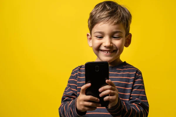 Front view portrait of small caucasian boy holding a smartphone in front of yellow background - little kid using mobile phone to take photos or play games while standing in studio with copy space