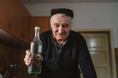 Front view portrait of senior caucasian man pensioner - real people elderly grandfather standing at home at farm looking to the camera in day holding bottle of homemade alcohol drink rakija brandy clipart