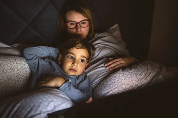 Mother and son watching movie or cartoon using laptop in bed at nighttime - Small boy and his mom using computer while lying in bed at night in dark room - leisure activity family bedtime concept