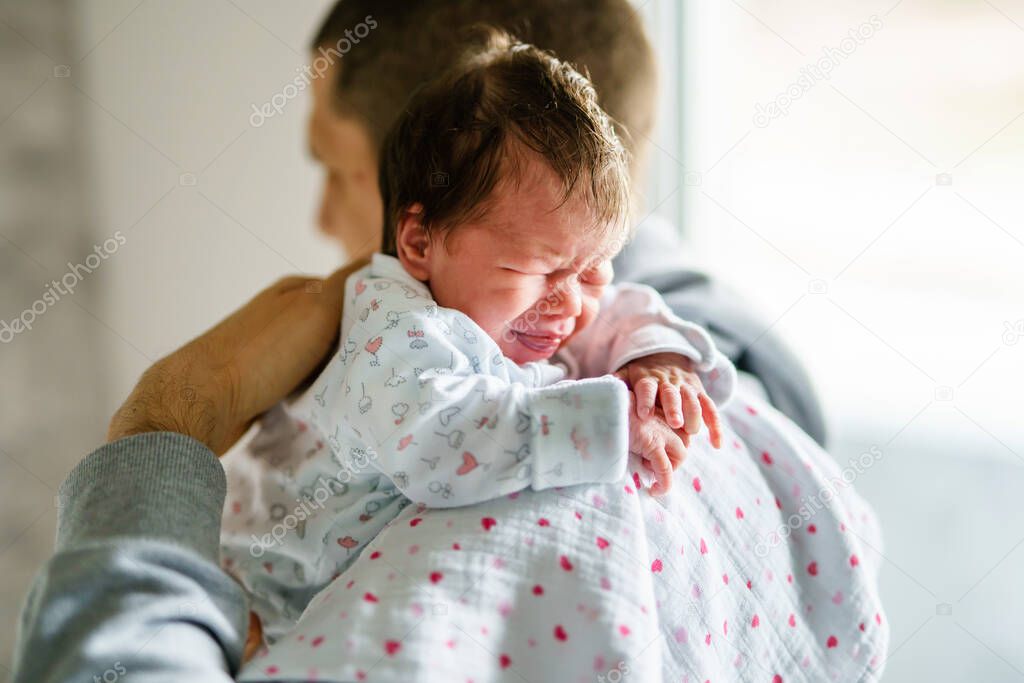 Back view of father holding his newborn baby over the shoulder while standing by the window in day at home - crying infant in position for burping making angry face new life and parenthood concept