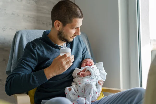 Caucasian man male father standing by the window at home holding and bottle feeding milk to his newborn baby daughter or son in day father's day parenthood real people concept