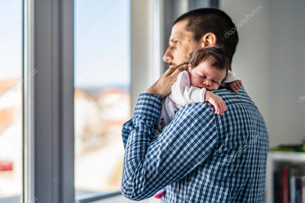 Side view of father holding his newborn baby over the shoulder while standing by the window in day at home - sleepy infant in position for burping new life and parenthood concept