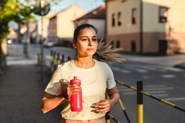 Ön manzara, şehrin açık havada kaldırımlarında koşan beyaz bir kadının yarı uzunluğunda güneşli bir günde spor antrenmanı ve eğlence anlayışı. Gerçek insanlar mekanı taklit eder.