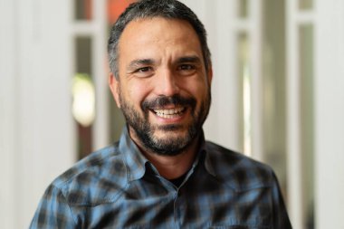 Sakallı, kısa saçlı, mutlu beyaz bir adamın resmi. Ofiste ya da evde tişört giymiş kameraya bakıyor.